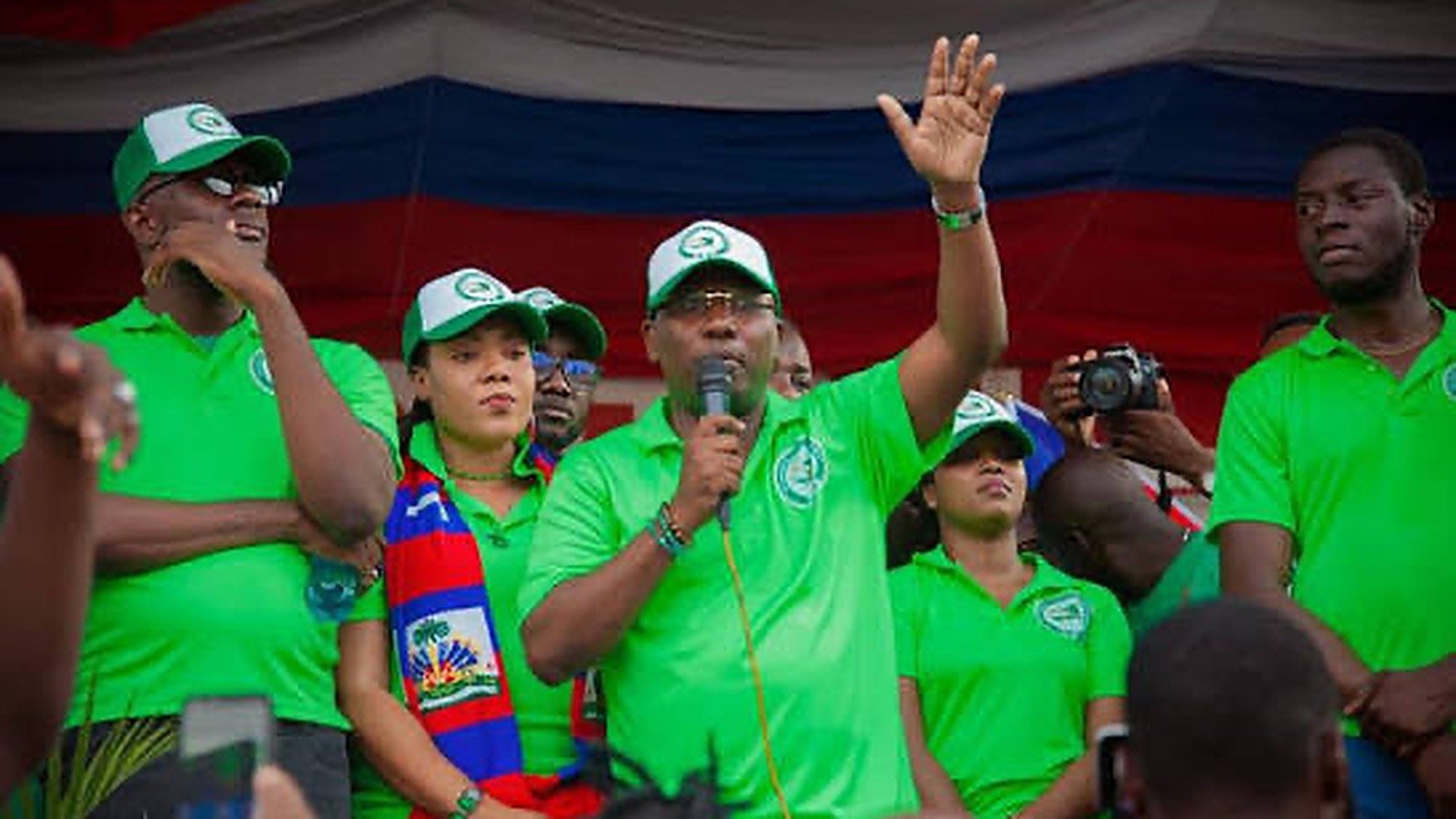 Claude Joseph, candidat du parti EDE à la présidentielle haïtienne, s'exprimant au micro lors d'un rassemblement de la formation, entouré de militants en tee-shirts verts EDE devant un drapeau haïtien géant.