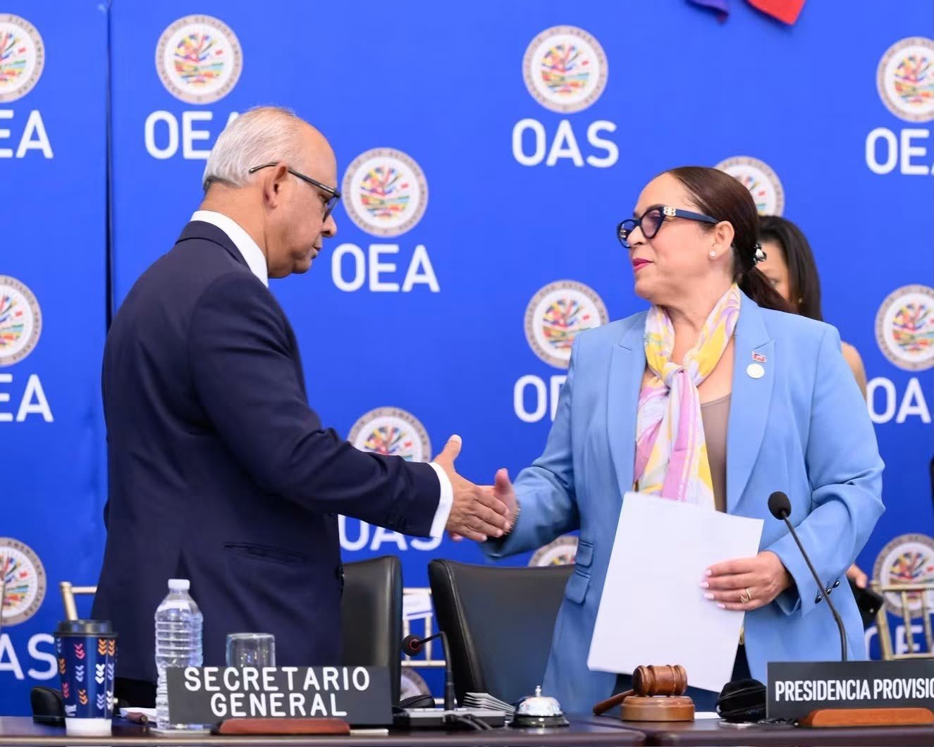 La ministre Raina Forbin, tailleur bleu ciel et foulard coloré, serre la main du secrétaire général de l'Organisation des États américains devant un mur d'images répétant les sigles « OEA » et « OAS », pancartes « Secretario General » et « Presidencia Provisional » posées sur la table.