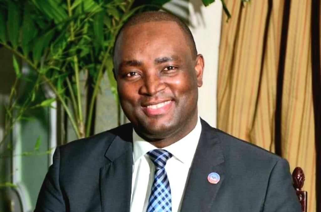 Portrait du diplomate haïtien Jean Tholbert Alexis, souriant, costume sombre et cravate, dans un intérieur.